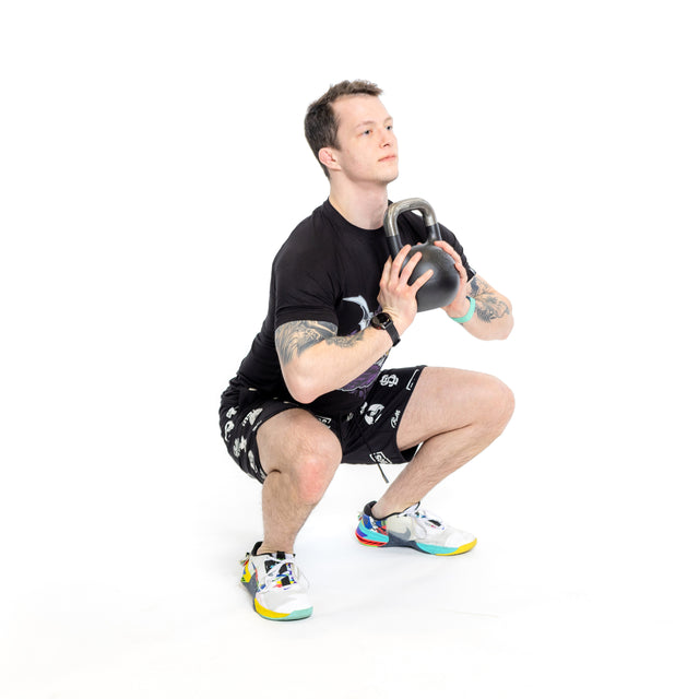 A person in Bells of Steel Gym Shorts, a black t-shirt, and colorful sneakers performs a squat with a kettlebell at chest height against a plain white background.
