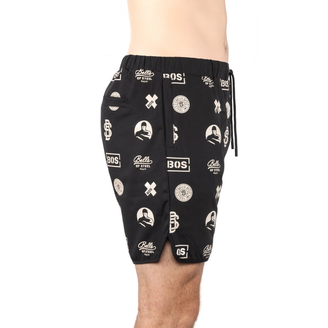 A person wears Bells of Steel Gym Shorts, featuring black stretchy fabric, white graphic prints, and a drawstring waistband, shown waist to above the knee against a plain white background.