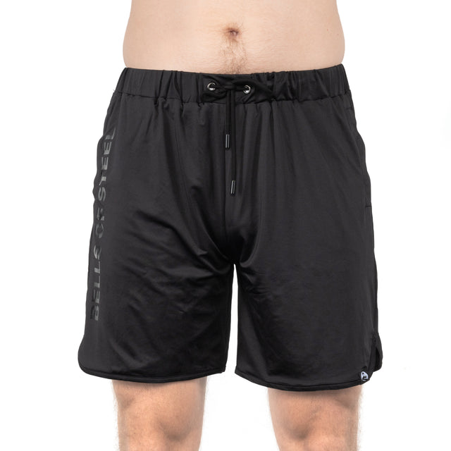 A person wears Bells of Steel Gym Shorts made from stretchy fabric with a drawstring waist and "BELLS OF STEEL" printed vertically on the left leg. Only their torso and upper legs are shown.
