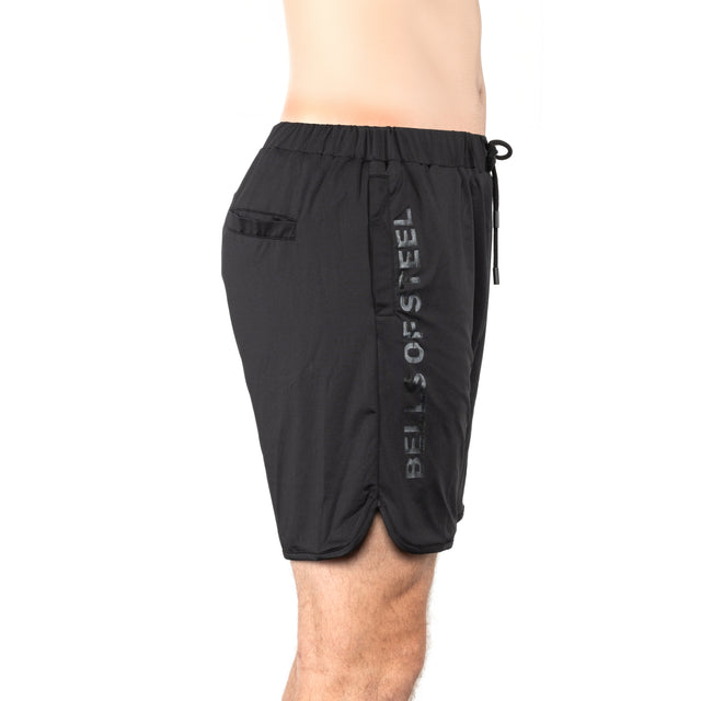 A person is shown from the side against a white background wearing Bells of Steel Gym Shorts in black, featuring "BELLS OF STEEL" vertically on the side, crafted from stretchy fabric with an elastic waistband and a small pocket.
