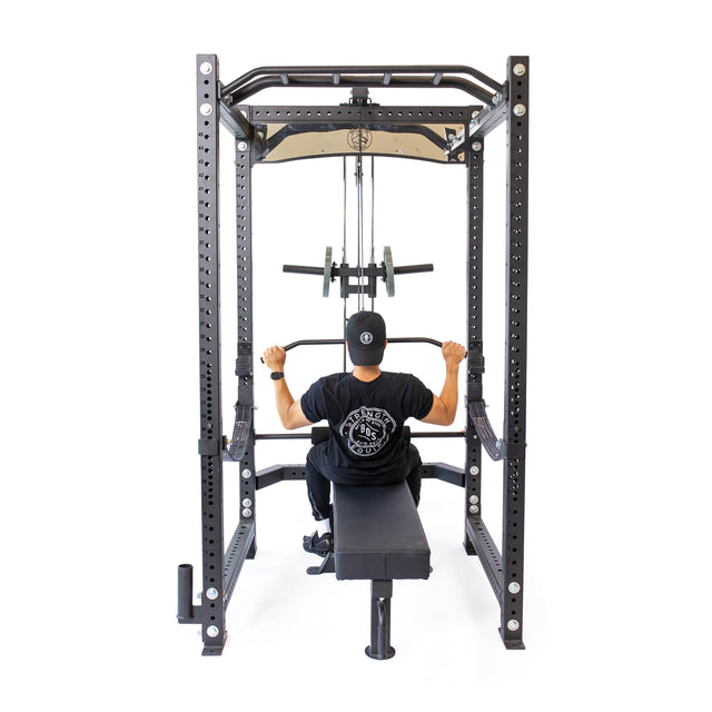 A person in a black shirt and hat uses the Bells of Steel Lat Pulldown & Low Row Rack Attachment—Hydra & Manticore—for lat pulldowns. Seated on a bench, gripping the bar with both hands, they're viewed from behind against a white background.