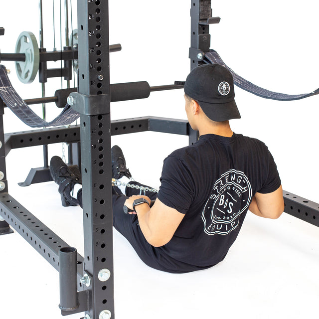 Wearing black athletic gear and a cap, a person sits on the gym floor, holding the Bells of Steel Lat Pulldown & Low Row Rack Attachment for Hydra & Manticore racks. Olympic Weight Plates and a power rack are visible nearby.