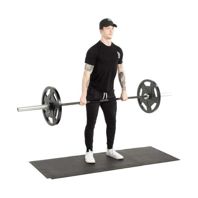 A person stands on Bells of Steel 24" x 24" Puzzle Mats (Set of 8), holding a loaded barbell, dressed in black t-shirt, pants, and cap, preparing to lift.