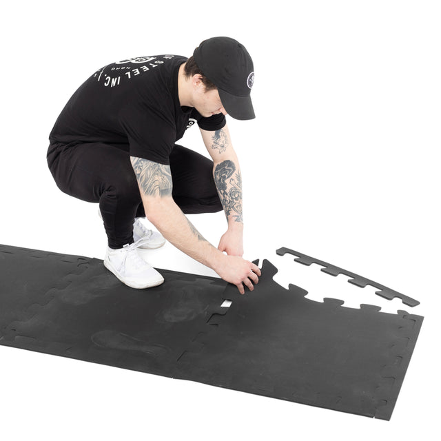 A person in black attire and a cap is kneeling on a white surface, assembling Bells of Steel 24" x 24" Puzzle Mats (Set of 8) to form gym flooring.