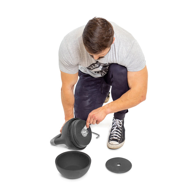 A person kneels on a white background assembling an adjustable dumbbell, surrounded by its parts and a Bells of Steel Adjustable Competition Kettlebell, both offering versatile weight options. They're dressed in a gray shirt and black pants.