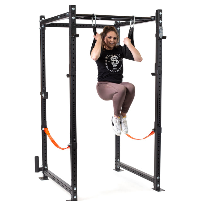 A woman in workout gear uses Bells of Steel Hanging Ab Straps (Pair) for arm support as she hangs from gymnastic rings on a power rack, knees bent and feet off the ground, smiling at the camera during her core workout.