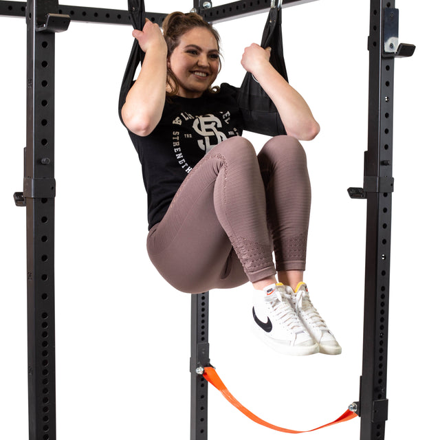 A woman in a black shirt, mauve leggings, and white Nike sneakers uses the Bells of Steel Hanging Ab Straps (Pair) attached to a pull-up bar for a core workout against a white background, smiling as she exercises.