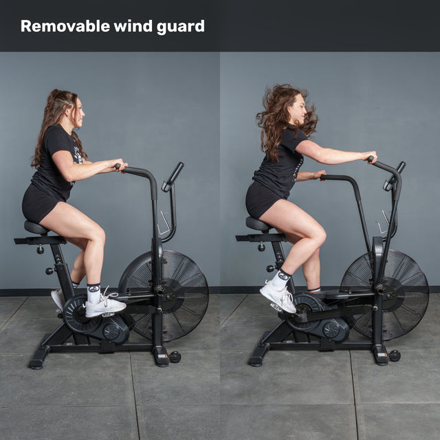 Two side-by-side photos show a woman in athletic attire riding the Bells of Steel Residential Air Bike with Wind Guard in a home gym, positioned against a gray wall.
