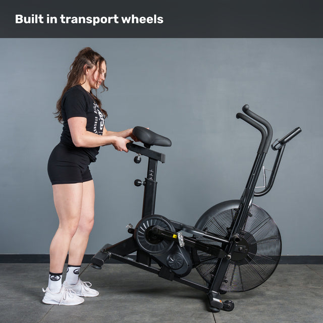 A woman in athletic wear tilts the Bells of Steel Residential Air Bike with Wind Guard onto its front wheels for easy moving. Above her, the caption reads, "Built in transport wheels." Shown indoors, it’s the ideal addition to any home gym.