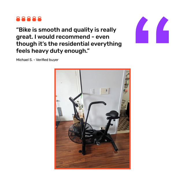 A Bells of Steel Residential Air Bike with Wind Guard sits on a wooden floor in a home gym. Verified buyer Michael S. praises the fan bike as smooth, high-quality, sturdy, and highly recommends it.