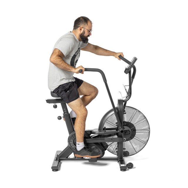 A man pedals on a Bells of Steel Residential Air Bike with Wind Guard, ideal for home gym cardio. Wearing a gray t-shirt and black shorts, he uses the bike's front fan wheel, handlebars, and digital display against a white backdrop.