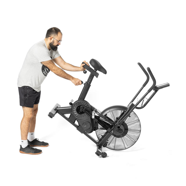 A person in athletic clothing adjusts a Bells of Steel Residential Air Bike with Wind Guard, featuring large fan-like wheels, ideal for home gym cardio workouts. The scene is set against a plain white background.