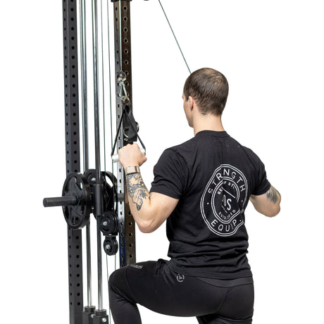 Wearing a black "Strength Equipment" t-shirt, a person kneels on one knee and performs a cable lat pulldown with Bells of Steel Aluminum Handles Pair. The gym background is white.