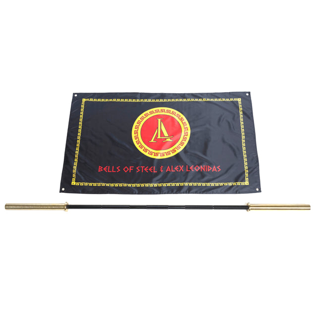 A black flag with a red and gold Greek-style emblem reading "BELLS OF STEEL & ALEX LEONIDAS" is displayed behind the Bells of Steel Alex Leonidas Onyx Bar, featuring gold sleeves and a sleek Cerakote finish.