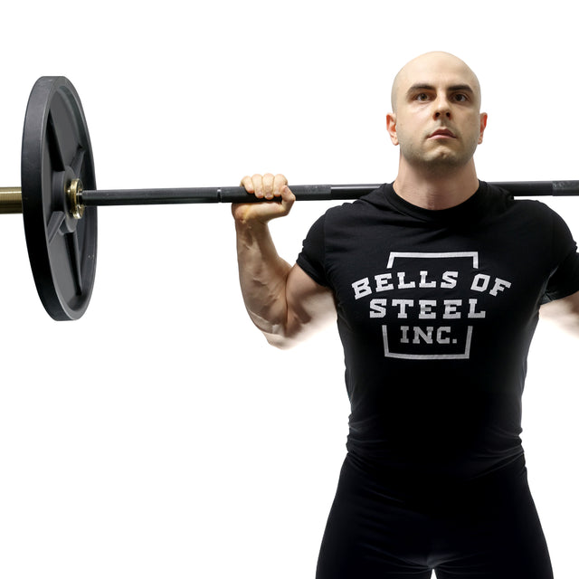 A muscular man in a "Bells of Steel" shirt lifts the Alex Leonidas Onyx Bar with heavy weights across his shoulders, standing against a white background.