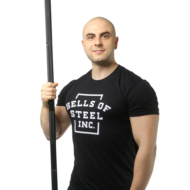 A muscular man in a Bells of Steel shirt holds the Alex Leonidas Onyx Bar—a premium power bar with a Cerakote finish—standing against a plain white background.