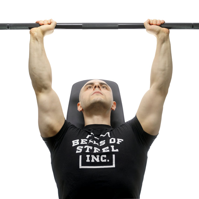 Wearing a black Bells of Steel shirt, a person lies on a bench pressing the Bells of Steel Alex Leonidas Onyx Bar, its Cerakote finish gleaming above their chest. The image is taken from a low angle.