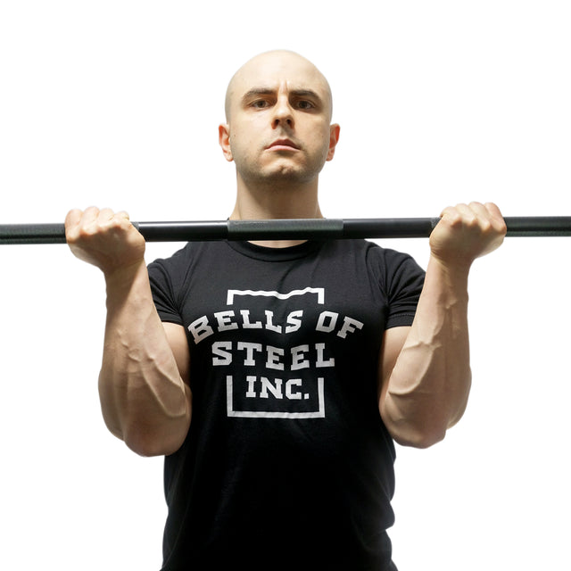 A man in a Bells of Steel shirt holds the Alex Leonidas Onyx Bar by Bells of Steel with both hands, palms up, curling the bar against a plain white background.