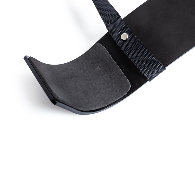 A close-up of the Bells of Steel Arm Blaster, a black shoehorn-shaped accessory with a padded, curved end and adjustable strap secured by a metal rivet—perfect for supporting proper bicep curl form—photographed on a white background.