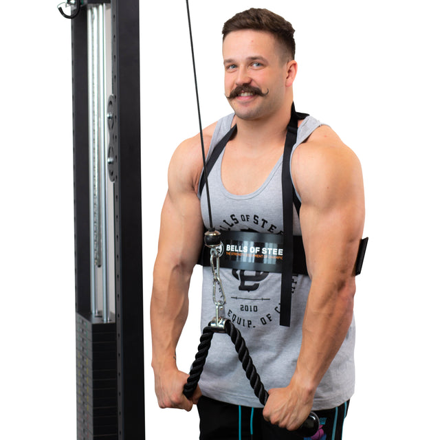 A man in a gray tank top uses the Bells of Steel Arm Blaster with a cable machine and rope attachment for triceps exercises, standing upright, smiling at the camera against a plain white background.