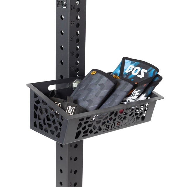 The Bells of Steel Rack Attached Storage Basket is a black metal organizer with a geometric cutout design and "BOS" logo, perfect for holding wrist wraps and gym essentials securely on your rack.