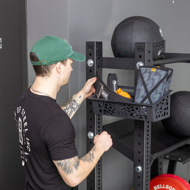 A person in a green cap and black t-shirt organizes workout gear on the Bells of Steel Rack Attached Storage Basket, a black metal shelf for medicine balls and fitness accessories.