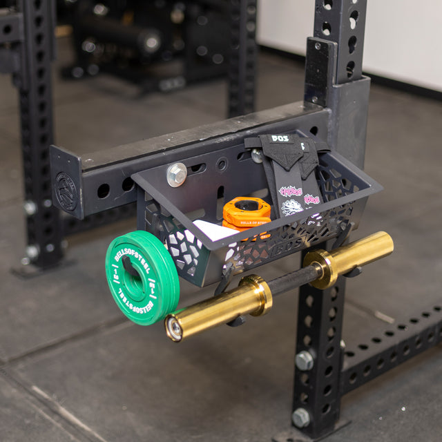 The Bells of Steel Rack Attached Storage Basket holds wrist wraps, chalk, tape, and stickers on a gym squat rack. Below it, a mini barbell with green plates is racked in a gym with black rubber flooring.