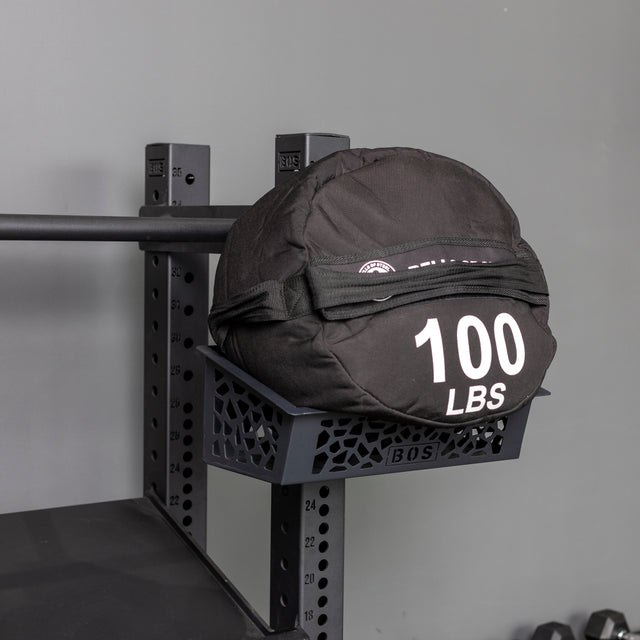 A Bells of Steel Rack Attached Storage Basket holds a black sandbag labeled "100 LBS" on a metal rack, set against a gray gym background.