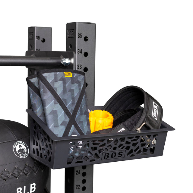 The Bells of Steel Rack Attached Storage Basket stores a patterned knee sleeve, black weightlifting belt, and yellow grip trainer as a gym organizer. A black medicine ball with white text rests beneath the basket.