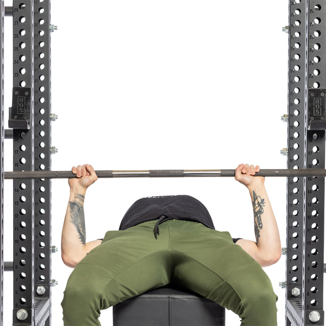 A person in green pants and a black shirt lies on a bench, gripping a barbell to bench press in a power rack equipped with Bells of Steel Standard J-Cups.