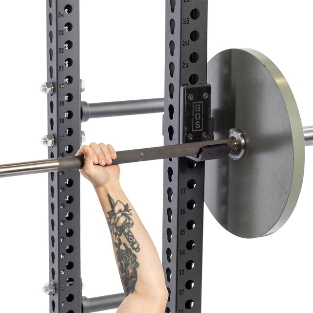 A tattooed arm lifts a barbell inside a black power rack with numbered height markings, using sturdy Bells of Steel Standard J-Cups that feature UHMW protection.