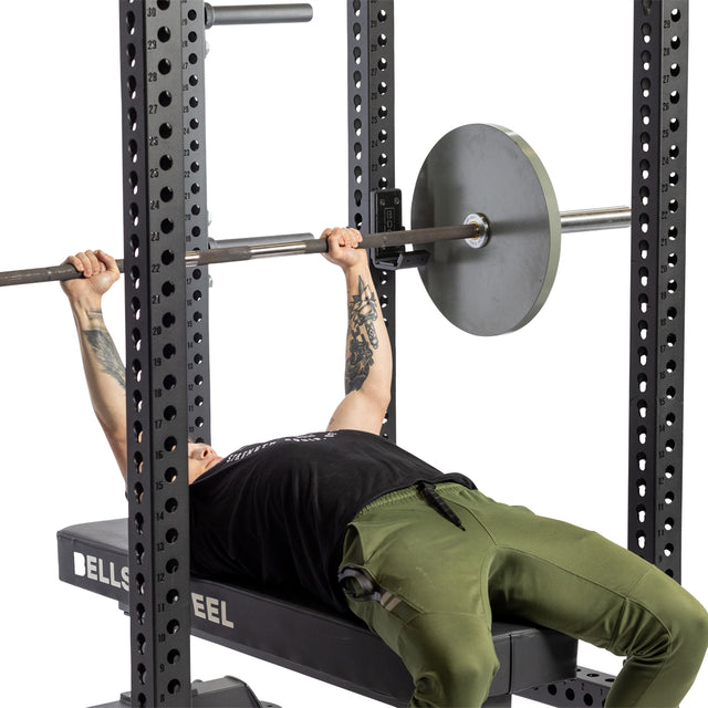 A person lies on a bench in a gym, gripping a barbell with weights, preparing to perform a bench press inside a power rack equipped with Bells of Steel Standard J-Cups.