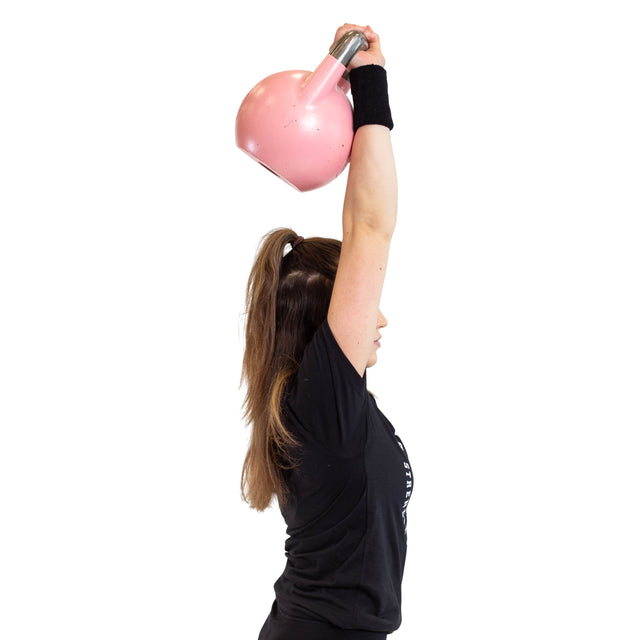 A woman in a black t-shirt wears Bells of Steel BellGuard Kettlebell Wrist Guards while lifting a large pink kettlebell overhead with one arm, her long brown hair in a ponytail, against a white background.