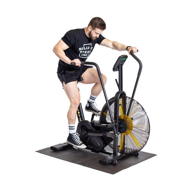 A man in a black t-shirt, shorts, and sneakers uses an air bike on a Bells of Steel Cardio Machine Floor Mat. He grips the handles as he pedals, with the plain white background emphasizing his workout on the premium mat.