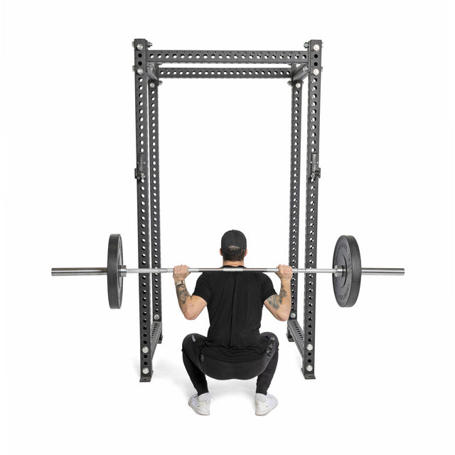 A person in black athletic wear and a cap squats with Bells of Steel All-Black Bumper Plates on a barbell inside a power rack, viewed from behind against a white background.