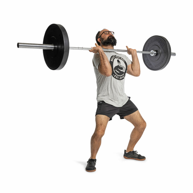 A man in glasses, a gray T-shirt, and shorts prepares to overhead press a barbell loaded with Bells of Steel All-Black Bumper Plate Sets. He stands with bent knees, looking up against a plain white background.