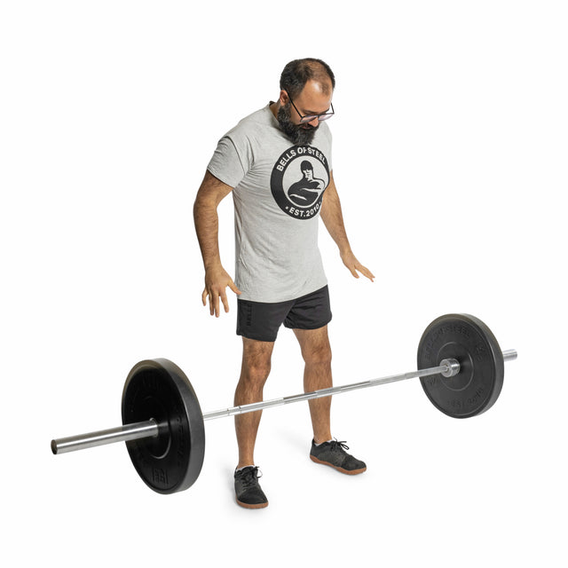 A man in a gray t-shirt and black shorts stands on a white background, preparing to lift a Bells of Steel All-Black Bumper Plate Set, focusing on safety as he looks down at the loaded barbell.