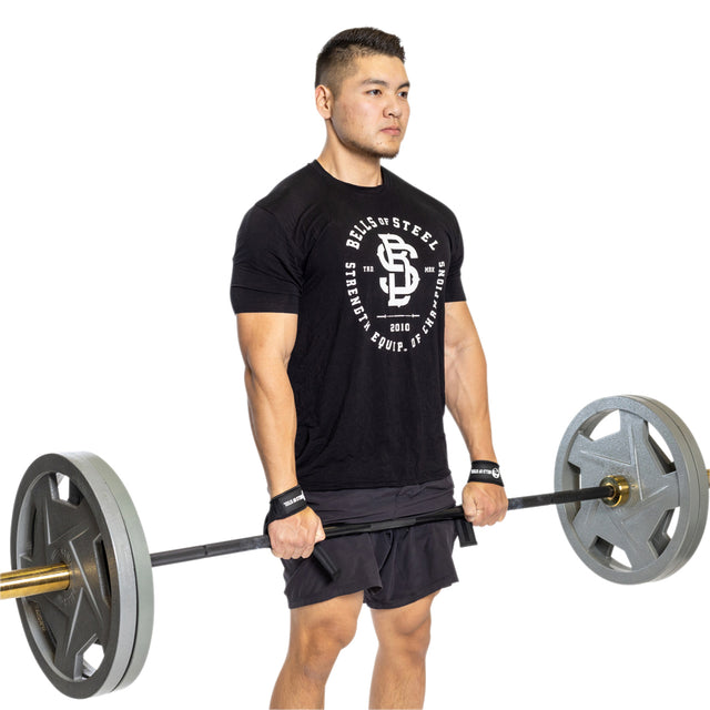A man wearing a black T-shirt and shorts uses Bells of Steel Lifting Straps to grip a loaded barbell at thigh height, preparing for a deadlift. He looks forward with focus against a white background.
