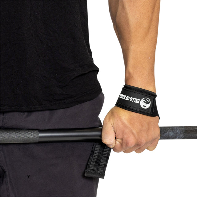 A person grips a barbell with one hand using Bells of Steel Lifting Straps, ready for strength training. They are dressed in a black shirt and dark shorts.