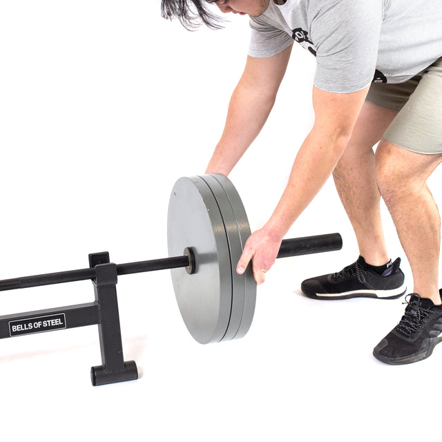 Wearing athletic gear, a person loads large weight plates onto a Bells of Steel Powerlifting Bar resting on a stand against a white background.
