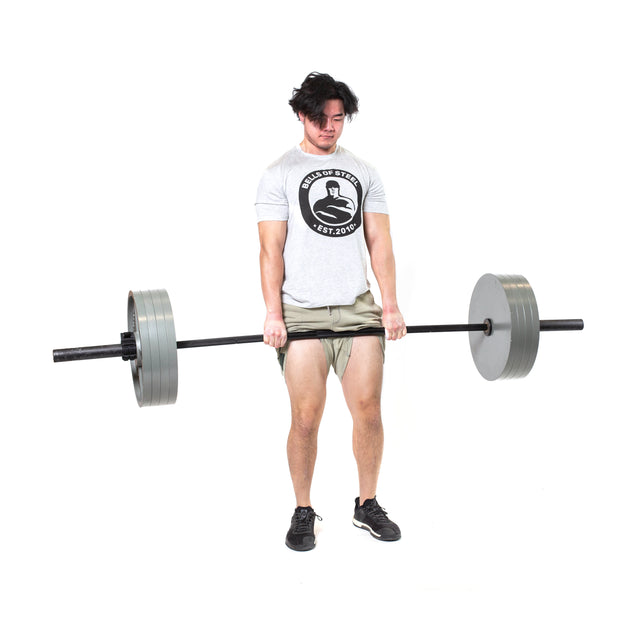 A man in a "Skills of Steel" t-shirt and shorts lifts the Bells of Steel Powerlifting Bar with large weights, standing against a plain white background.