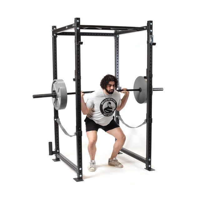 A man in a white T-shirt and black shorts performs a barbell squat inside a black power rack, using the Bells of Steel Powerlifting Bar loaded with weight plates.