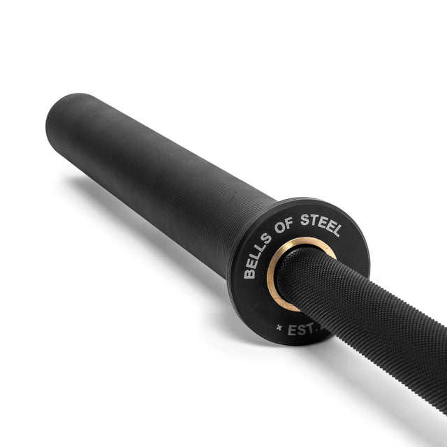 A black Bells of Steel Powerlifting Bar with "BELLS OF STEEL * EST." on the end cap lies diagonally on a white background.