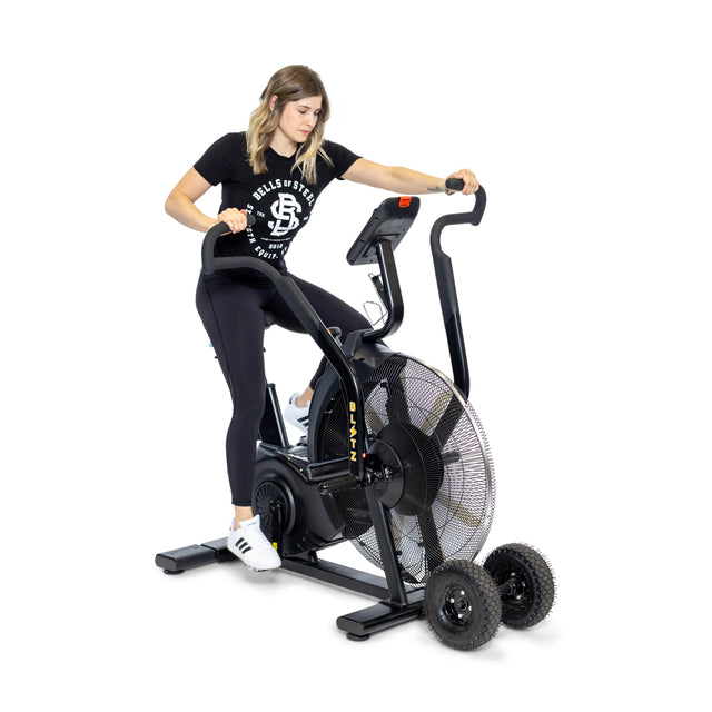 A woman in athletic wear sits on the Bells of Steel Blitz Max Air Bike, gripping the handlebars, with two replacement wheels on the floor beside this commercial fitness equipment against a plain white background.
