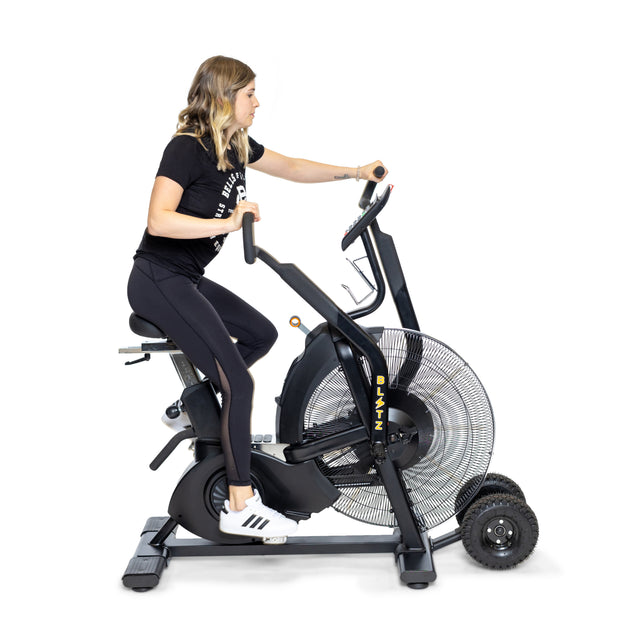A woman in athletic wear pedals and grips the handles of a Bells of Steel Blitz Max Air Bike against a plain white background.