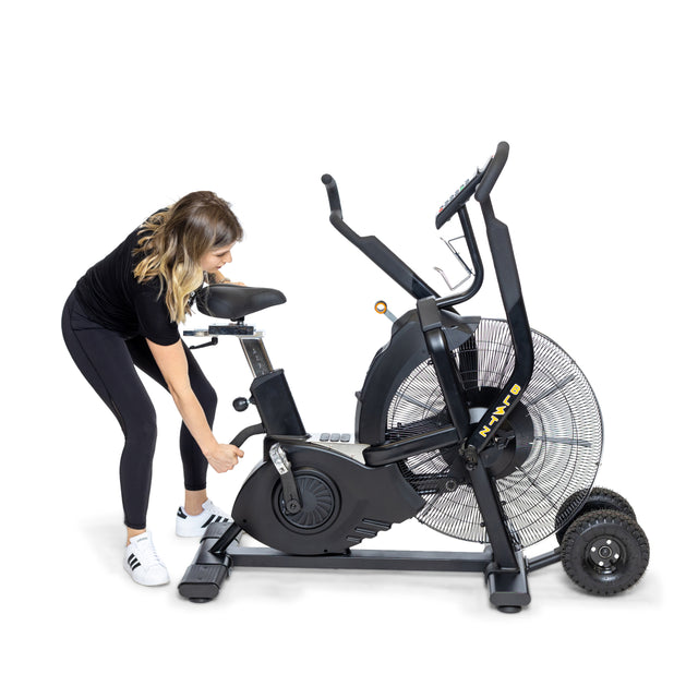 A woman in athletic wear adjusts the seat of a Bells of Steel Blitz Max Air Bike, a commercial fitness staple, against a white background.