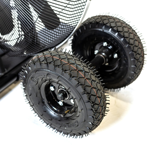 Close-up of two rugged, black rubber wheels with deep treads on a metal axle, part of the Bells of Steel Blitz Max Air Bike, with a mesh guard visible in the upper left corner against a white background.