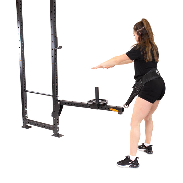 Wearing a black outfit, someone uses the Bells of Steel Belt Squat / Lever Arms Rack Attachment on a metal frame with a harness for resistance training, perfectly complementing their home gym's white, minimalistic backdrop.