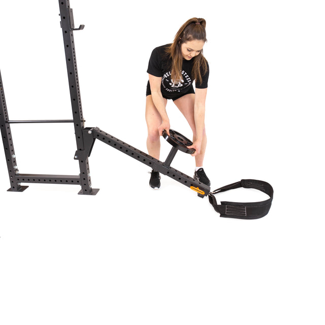 Dressed in a black T-shirt and shorts, a person sets up a sleek home gym with the Bells of Steel Belt Squat / Lever Arms Rack Attachment. Holding a weight plate, they adjust the black strap on the attachment against a plain white backdrop, preparing to enhance their workout routine.