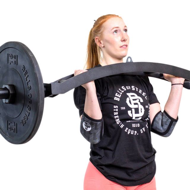 A light-skinned, red-haired woman lifts a curved barbell while wearing pink leggings, a black Bells of Steel shirt, and Bells of Steel Elbow Sleeves for support. The background is white.